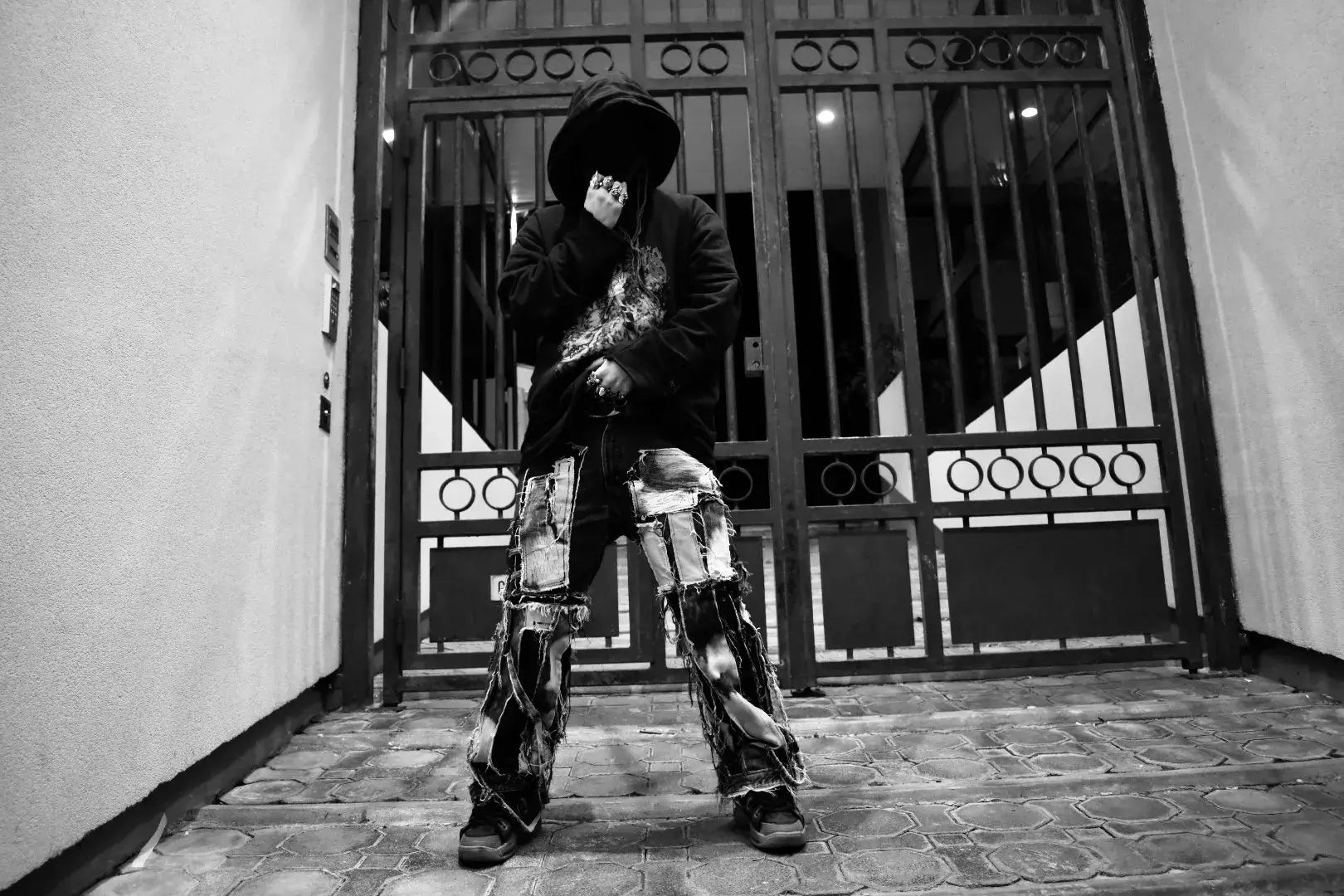 Masked model posing in front of a Parisian gate, wearing destructured denim and oversized hoodie from PrototypesParis ConceptWear line.