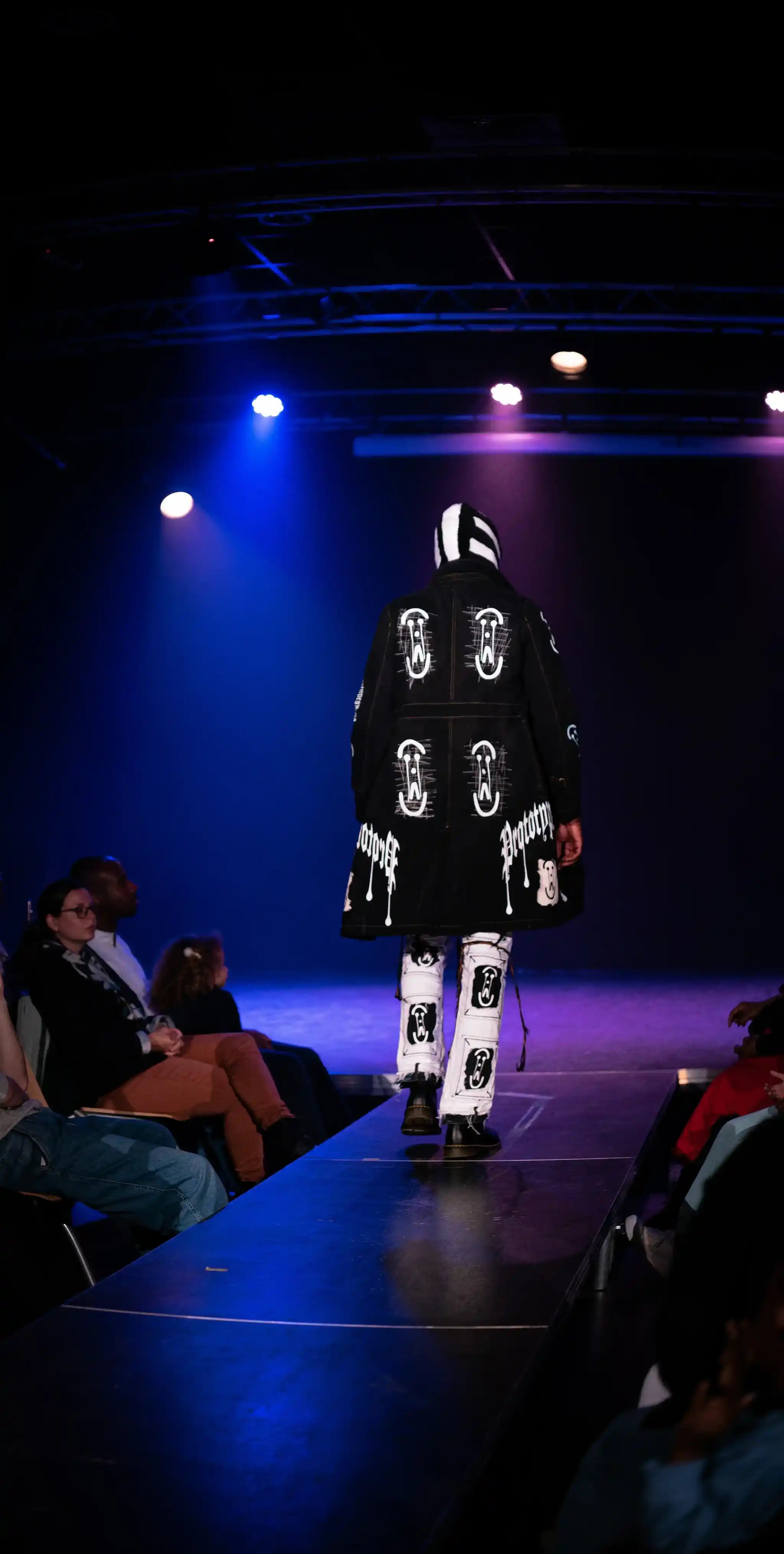 Runway photo: PrototypesParis graphic black trench with white logo patches over printed pants—rear view under stage lights.
